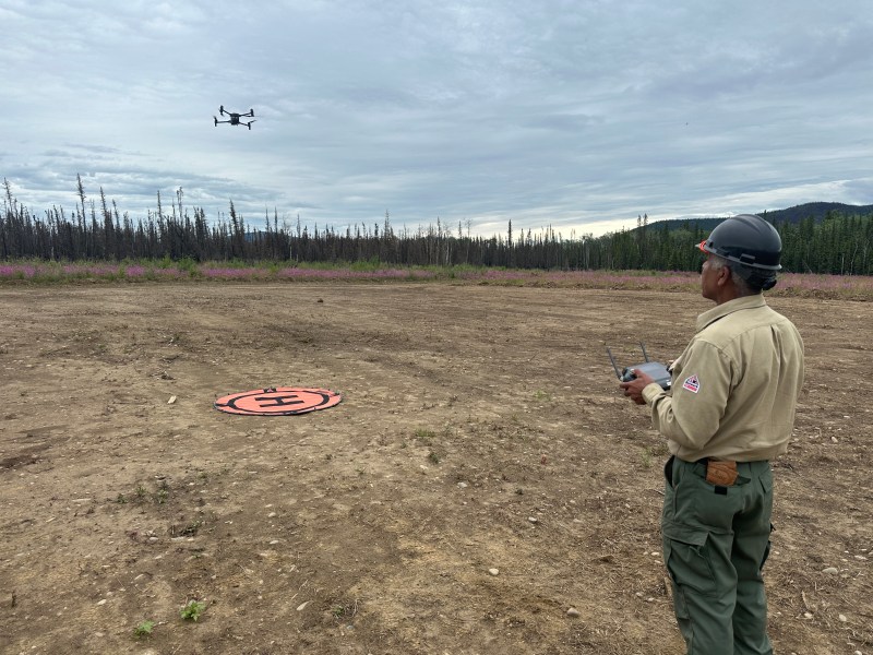 A man in a firefighting unifrom operates a drone over a cleared area on the perimeter of a forest fire