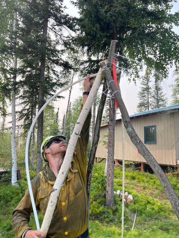 A firefighter reaches up into a tripod made of cut branches in order to position a hose to spray water.