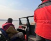 A firefighter with a life jacket sits in a boat that is moving along the Yukon River. One person stands in an orange jacket to the right of the image, steering the boat.