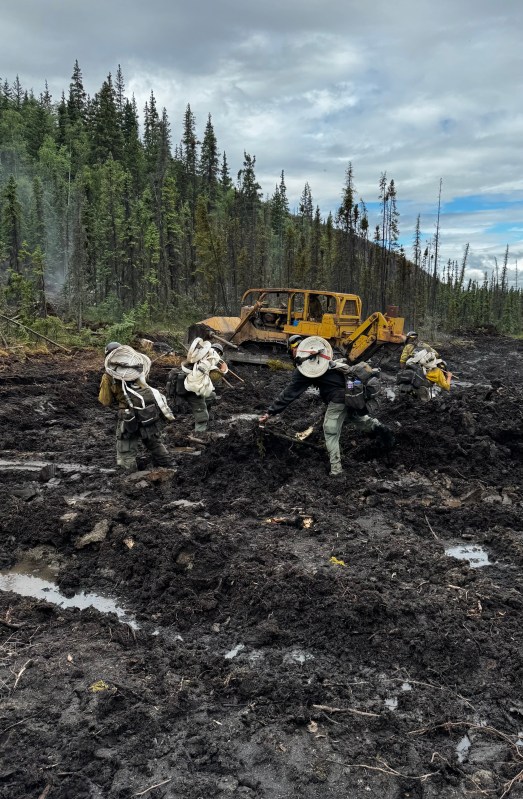 Firefighters clearing vegetation and working in a muddy area near heavy machinery in a forested landscape.