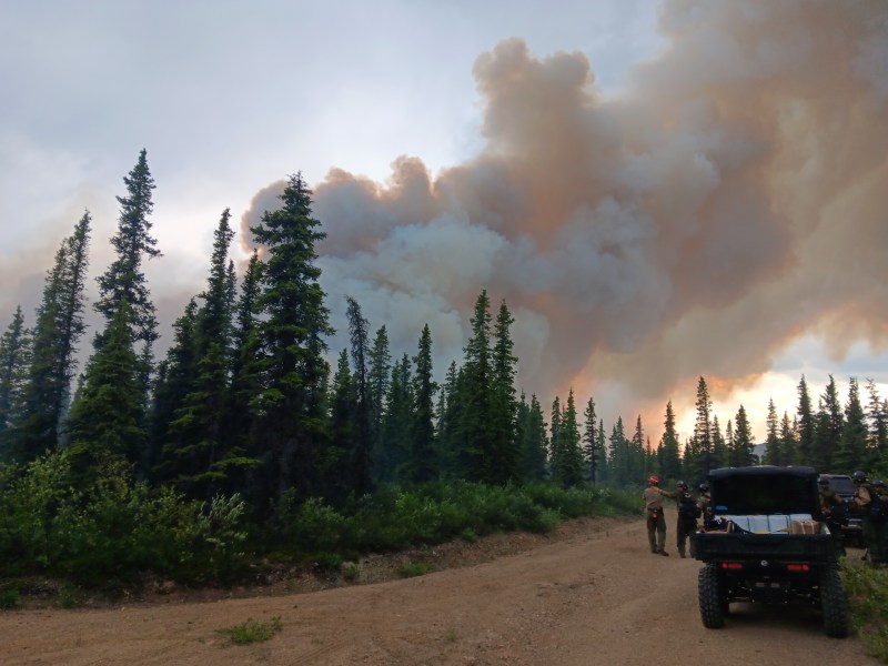 Large smoke plume behind trees, firefighters and ATV in the foreground