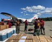 Eight members of the Midnight Suns Hotshot crew are standing next to a small white plane with a red stripe. They have unloaded supplies from the plane and are waiting to be transported off of the fire after working two weeks. They are all wearing burgundy crew shirts and green Nomex pants. In the front of the photo, stacks of supplies are visible - cardboard boxes of supplies and blue and white coolers. The sky above is mostly blue with scattered white clouds.