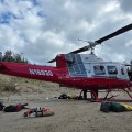 A red and white helicopter sits on a sand and gravel with various equipment such as a chainsaw, tools and bags lying on the ground near the helicopter.