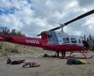 A red and white helicopter sits on a sand and gravel with various equipment such as a chainsaw, tools and bags lying on the ground near the helicopter.