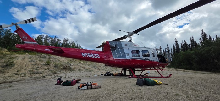 A red and white helicopter sits on a sand and gravel with various equipment such as a chainsaw, tools and bags lying on the ground near the helicopter.