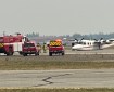 An aircraft sits without landing gear on a runway with numerous red emergency response vehicles parked in front of it.