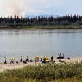 A group of wildland firefighters dressed in yellow and green Nomex stand on the bank of a large river next to a boat. Wildfire smoke rises from the forest on the opposite river bank.