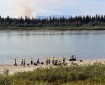 A group of wildland firefighters dressed in yellow and green Nomex stand on the bank of a large river next to a boat. Wildfire smoke rises from the forest on the opposite river bank.