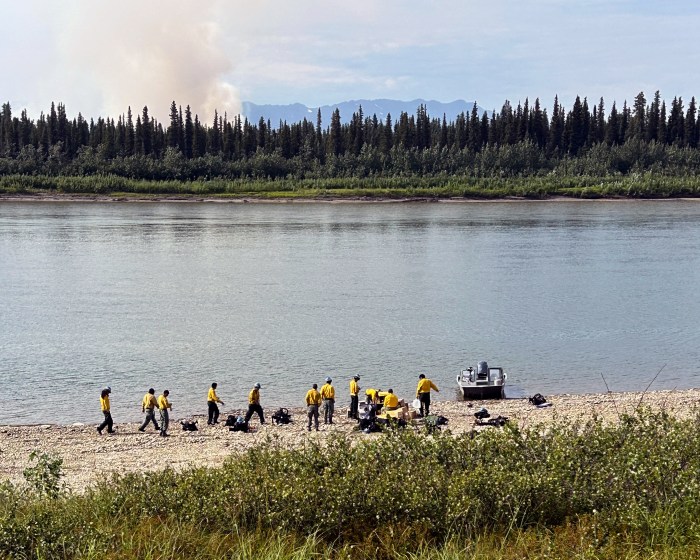 A group of wildland firefighters dressed in yellow and green Nomex stand on the bank of a large river next to a boat. Wildfire smoke rises from the forest on the opposite river bank.