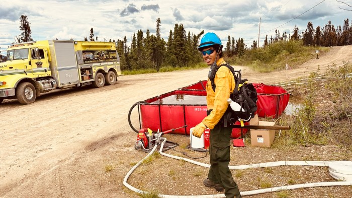 A firefighter in full Nomex, and hard hat and gloves walks past a red collapsible tank with pump and hose attached. A large yellow water tender is parked across a dirt road from him.