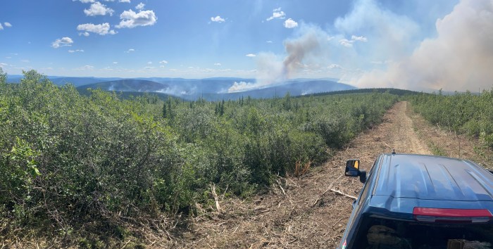 A wide area of smoke drfits and lifts skyward out of a forestsed area, as seen from the back of a truck