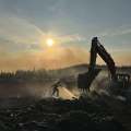 A firefighter douses a hot spot working in tandem with an excavator in the late afternoon sun and smoke filled sky
