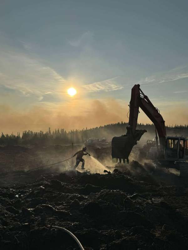 A firefighter douses a hot spot working in tandem with an excavator in the late afternoon sun and smoke filled sky