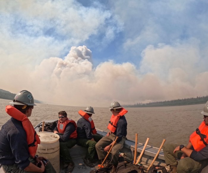 Firefighters with orange life vests sit in boat on the water with hand tools beside them. In the background a plume of smoke from the Lush fire fills a blue sky with light clouds.