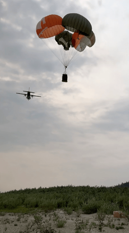 Supplies connected to a parachute drop from the sky as a plane flies away.