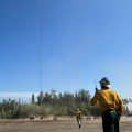 The photo shows a man wearing firefighter protective gear standing in the foreground with a helicopter lifting a load. In the midground a horizon of trees can be seen against a blue sky.