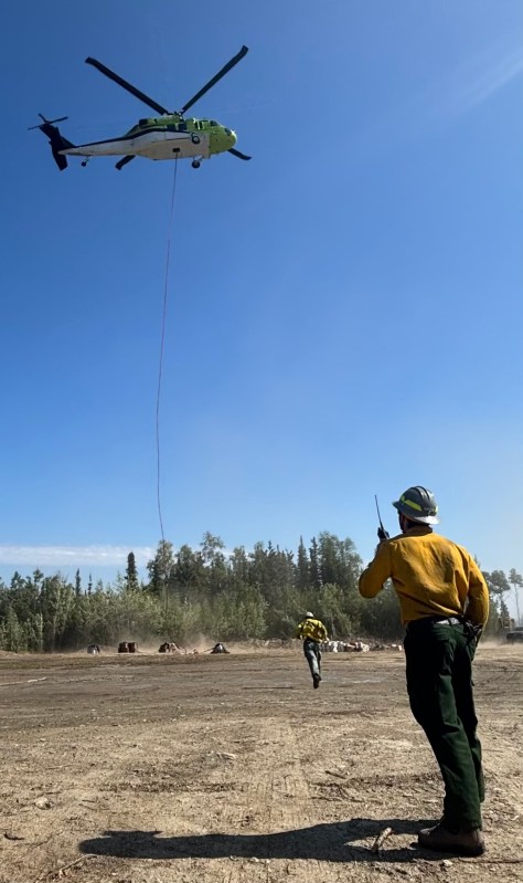 The photo shows a man wearing firefighter protective gear standing in the foreground with a helicopter lifting a load. In the midground a horizon of trees can be seen against a blue sky.