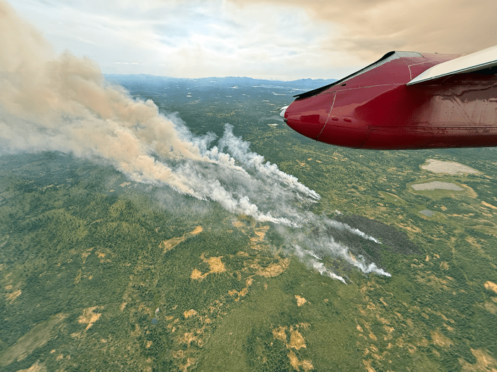 Smoke billowing up from a burned patch of green tundra. A red part of an aircraft wing is in the corner.