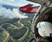 A white-helmeted person sits in the open door of an airplane looking at a landscape with smoke billowing up from a fire.
