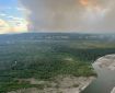 A wall of smoke rises up from a forest in the distance next to a river.