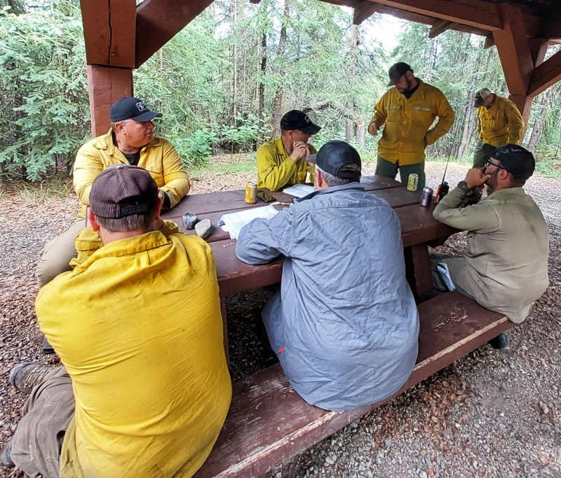 a group of firefighters gather around a picnic table covered with maps and radios