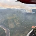 Looking out the spotters window of a smokejumper plane, a column of smoke rises in the distance from a wildfire. To the left of the frame, the Fortymile River flows, separated from the smoke column by one and a half miles of black spruce forest.