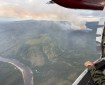 Looking out the spotters window of a smokejumper plane, a column of smoke rises in the distance from a wildfire. To the left of the frame, the Fortymile River flows, separated from the smoke column by one and a half miles of black spruce forest.