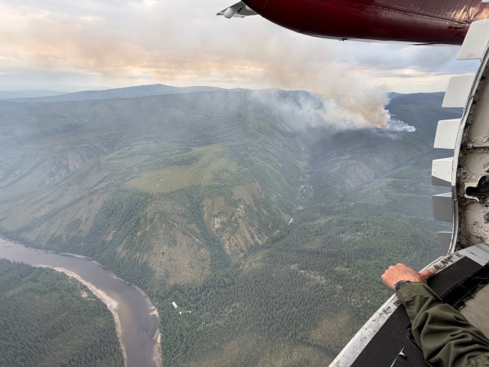 Looking out the spotters window of a smokejumper plane, a column of smoke rises in the distance from a wildfire. To the left of the frame, the Fortymile River flows, separated from the smoke column by one and a half miles of black spruce forest.