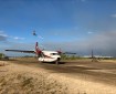 Image shows a rural dirt-surface airstrip with a landed plane in the foreground and a helicopter flying in the background. The skies are blue, but hazy with clouds and smoke.