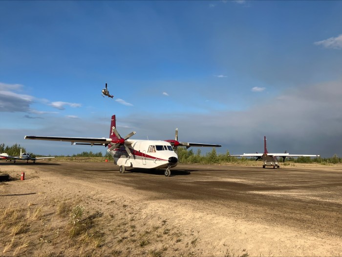 Image shows a rural dirt-surface airstrip with a landed plane in the foreground and a helicopter flying in the background. The skies are blue, but hazy with clouds and smoke.