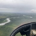 The inside view from the right hand side of a helicopter cockpit. Rain is visible on the glass cover, partially obscuring the view. In the distance, a river meanders through mixed timber.