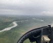 The inside view from the right hand side of a helicopter cockpit. Rain is visible on the glass cover, partially obscuring the view. In the distance, a river meanders through mixed timber.