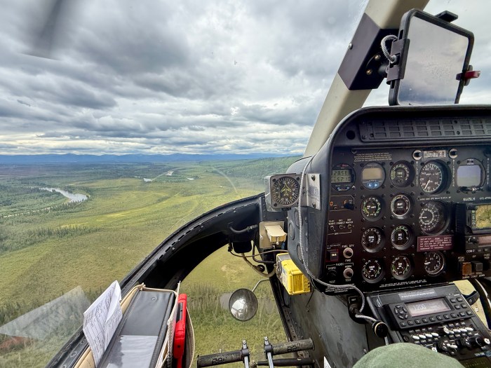 The inside of an aircraft cockpit with various gauges and reading instruments as it flies above vast green vegetation. A sliver of gray river can be seen in the photo background and the sky is overcast.