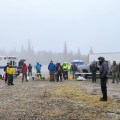 A man stands in the foreground wearing a hoodie and holding a microphone. He is speaking to a crowd of firefighters who are mostly dressed in hoodies and rainhgear. The ground is rocky and damp. Trees line the horizon of the photo. The trees appear as a silhouette in foggy conditions.