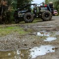 A water puddle on a gravel road is in the foreground. Trees are on both sides of the photo. A black skidgen is parked in the background with yellow flames painted on it.