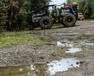 A water puddle on a gravel road is in the foreground. Trees are on both sides of the photo. A black skidgen is parked in the background with yellow flames painted on it.