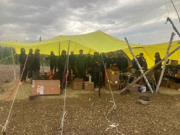 A group of 17 firefighters stand underneath a yellow tarp. The tarp is supported by wood posts and tied to the ground. The tarp covers most of the camp supplies for the firefighters.