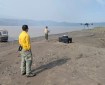 An Unmanned Aircraft System (UAS) comes in for a landing along the Yukon River. Two fire personnel are shown standing along the river with their yellow Nomex shirts and green Nomex pants. Across the river, an island with tall spruce is seen. The air is very smokey.