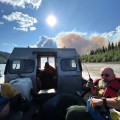This is a close up photo of the back of an open boat that is driving across the Yukon River. The boats are critical to shuttling people and equipment across the river from the Rampart airstrip to the fire area. One firefighter is in the back of the boat with protective gear on, including a red life vest. He is holding a radio for communication. They are going toward the fire, a white plume of smoke is visible above the boat, they are driving towards it. The sky is blue with a bright sun in the middle and white clouds in the upper left.