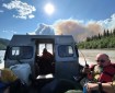 This is a close up photo of the back of an open boat that is driving across the Yukon River. The boats are critical to shuttling people and equipment across the river from the Rampart airstrip to the fire area. One firefighter is in the back of the boat with protective gear on, including a red life vest. He is holding a radio for communication. They are going toward the fire, a white plume of smoke is visible above the boat, they are driving towards it. The sky is blue with a bright sun in the middle and white clouds in the upper left.
