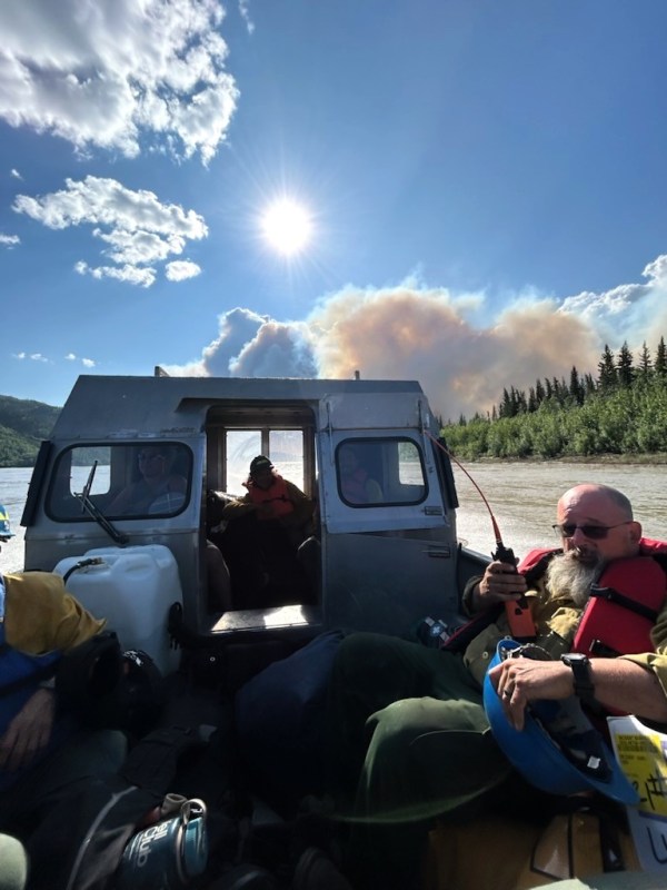 This is a close up photo of the back of an open boat that is driving across the Yukon River. The boats are critical to shuttling people and equipment across the river from the Rampart airstrip to the fire area. One firefighter is in the back of the boat with protective gear on, including a red life vest. He is holding a radio for communication. They are going toward the fire, a white plume of smoke is visible above the boat, they are driving towards it. The sky is blue with a bright sun in the middle and white clouds in the upper left.