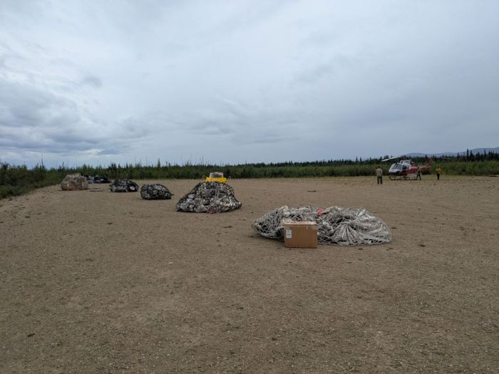 The bottom half of the image is bare, light brown earth with six netted piles of gear including white hose laid out. A helicopter is stationed further away on the bare dirt, with three firefighters nearby. There are cloudy blue-gray skies above the people and fire gear.