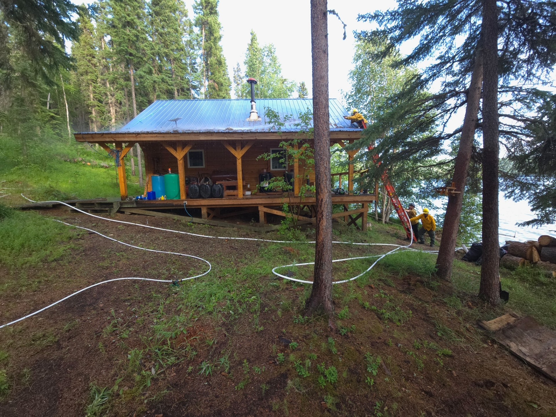Image of cabin with hose running along the front of it. Firefighters climb a ladder to continue laying hose around the structure