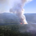 Aerial view of a wildfire burning in a dense forested area, with bright orange flames visible at the base of a tall, vertical smoke column rising into a clear blue sky. Surrounding the fire are rolling hills and a small lake to the right, with smoke drifting across the landscape.