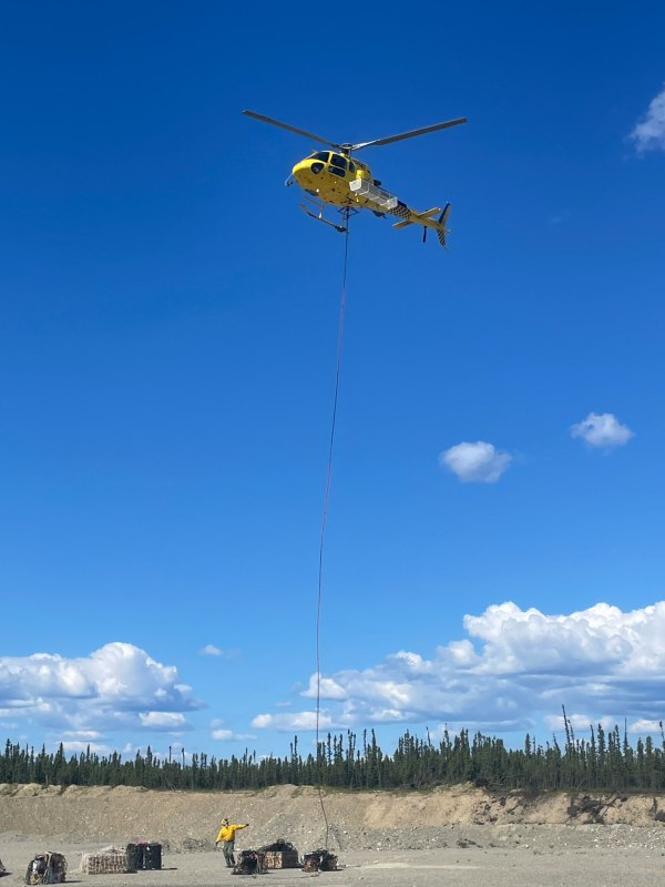yellow helicopter hovers over rocky landscape as a firefighter in a yellow shirt and green pants connects a long line to cargo in nets.