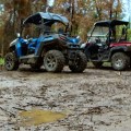 Two UTVs are parked near a wooded area that has recently been charred by fire. The UTV on the left is blue and black, while the one on the right is red and black. The ground is muddy and brown. A puddle of muddy water is the foreground and focus of the photo.