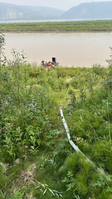 A hose stretches towards the Yukon River. A Mark 3 pump lies along the bank of the river, siphoning water. Knee high brush and grass cover the area.