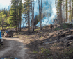 A group of firefighters works near the edge of a the 7 Mile Fire. Smoke rises from burning piles of woody debris. Fire vehicles are parked along a dirt road in the foreground, while flames and smoke are visible among the trees in the background.