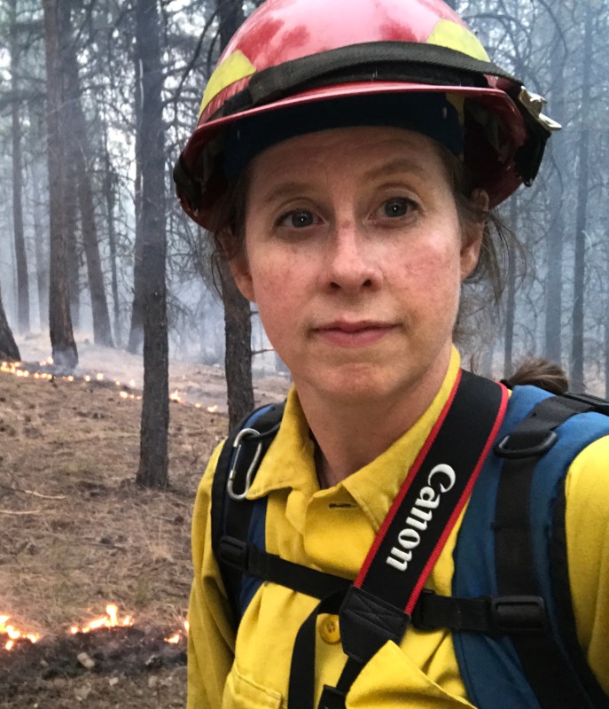 A woman wearing a red hard hat and a yellow shirt with a Canon camera strap around her neck.