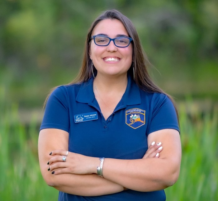 A smiling woman with long brown hair, a blue shirt with a name tag and blue eyeglasses.